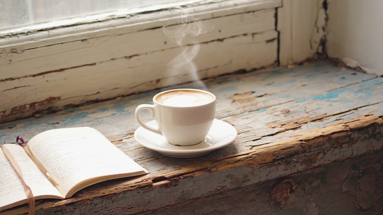Eine dampfende Tasse Kaffee steht im Morgenlicht auf einer hölzernen Fensterbank neben einem aufgeschlagenen Notizbuch.