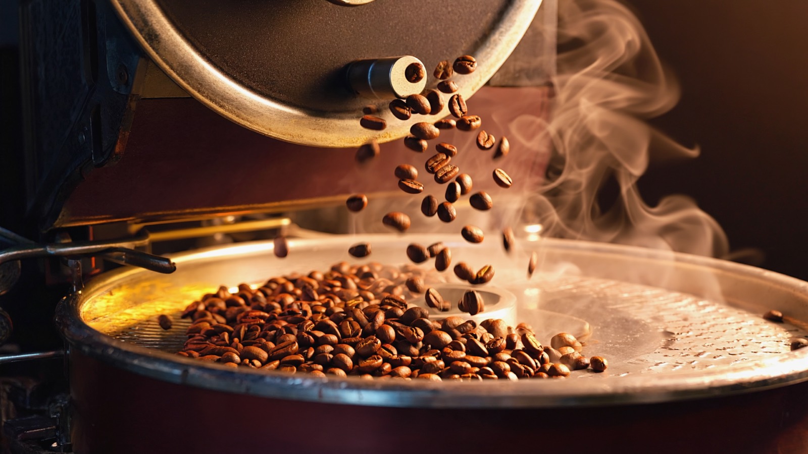 Rotbraune Kaffeebohnen fallen aus der Trommel eines Trommelrösters in das drehende Kühlsieb, Rauch steigt auf.